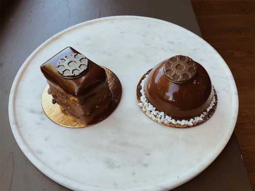 Two chocolate desserts on a marble plate: a square cake and a dome-shaped treat, both with decorative metallic accents.