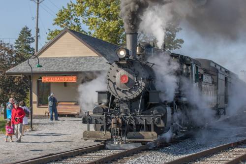 A steam locomotive billows smoke at a train station, with people nearby and a sign reading "Hollidaysburg."