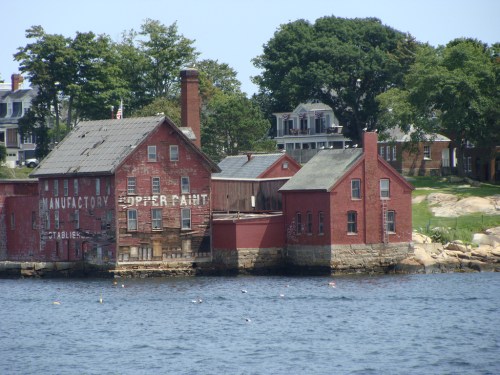 Historic red brick buildings by the water, featuring faded signage and surrounded by trees and houses.