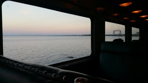 View from a train window overlooking a calm lake at sunset, with distant city structures visible on the shore.