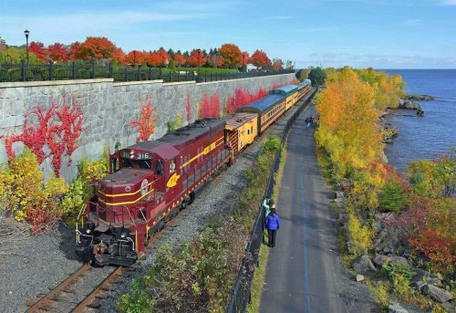 A colorful train travels along a scenic route by a lake, surrounded by vibrant autumn foliage and a walking path.