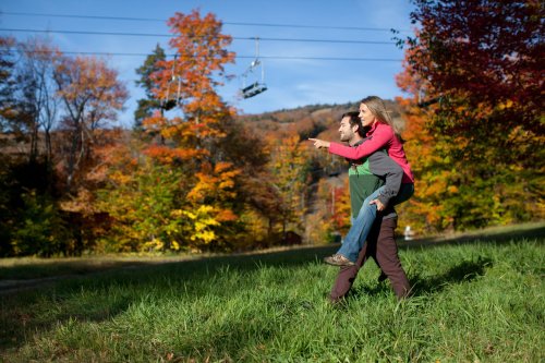 A couple enjoys a playful moment outdoors, surrounded by vibrant autumn foliage and a ski lift in the background.