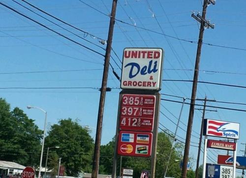 Sign for United Deli & Grocery displaying gas prices: $3.85, $3.97, $4.12, with nearby trees and power lines.