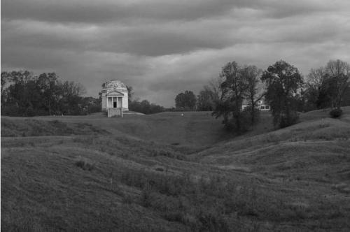 A black and white landscape featuring a small monument on a hill, surrounded by rolling grassy terrain and trees under a cloudy sky.