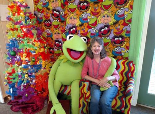 A woman sits beside a large Kermit the Frog plush in a colorful room decorated with a vibrant Christmas tree and Muppet-themed backdrop.