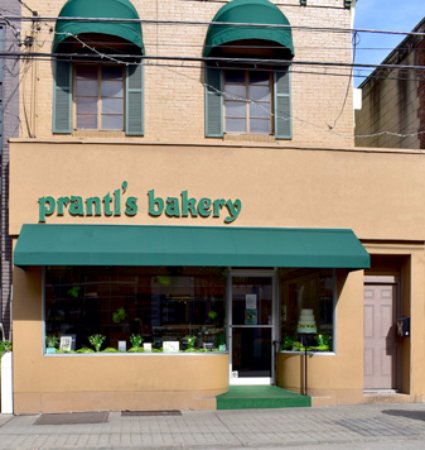 A storefront of Pranti's Bakery featuring green awnings and plants in the window, set in a brick building.