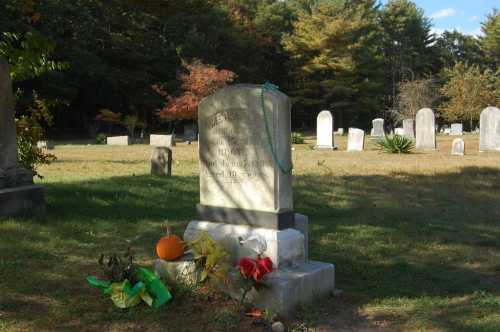 A gravestone in a cemetery surrounded by trees, with flowers and a small pumpkin placed at its base.