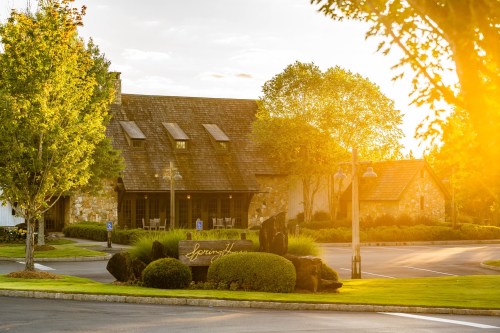 A charming stone building with a sloped roof, surrounded by trees and greenery, illuminated by warm sunset light.