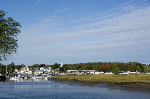 A serene waterfront scene with boats docked, surrounded by trees and buildings under a clear blue sky.