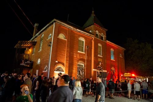 A crowd gathers outside a historic brick building at night, illuminated by orange lights and a fire truck nearby.