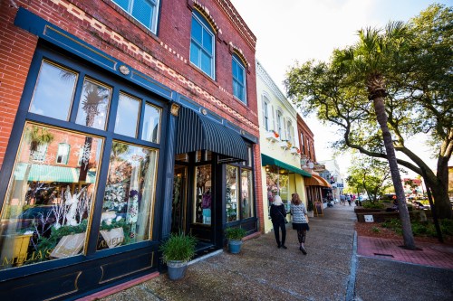 A charming street scene with brick buildings, shops, and people walking along a sunny sidewalk lined with greenery.
