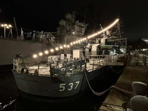 A navy ship docked at night, illuminated by string lights, with mooring lines and equipment visible on the deck.