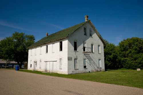 A weathered white building with a green roof, surrounded by grass and trees, under a clear blue sky.