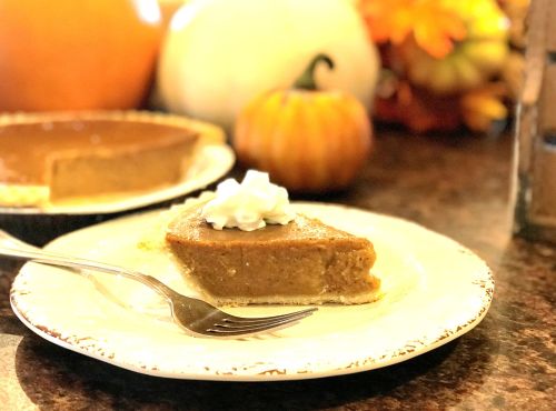 A slice of pumpkin pie topped with whipped cream on a decorative plate, with pumpkins and autumn decor in the background.