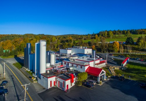 Aerial view of a large industrial facility surrounded by trees and a clear blue sky, with silos and a red-roofed building.
