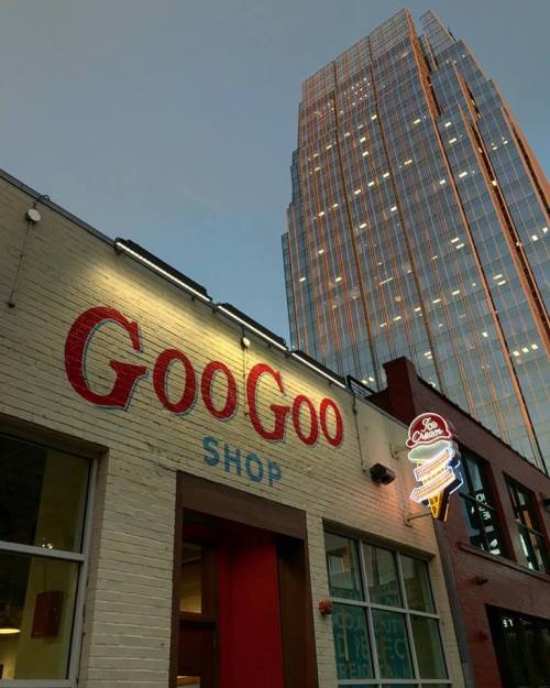 A brick building with "GooGoo Shop" sign, illuminated neon, and a tall modern skyscraper in the background at dusk.