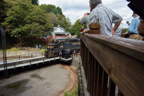 A train on a turntable with spectators watching from a wooden deck in a scenic outdoor setting.