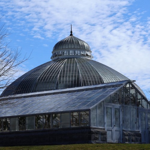 A large glass greenhouse with a domed roof, surrounded by blue sky and wispy clouds.