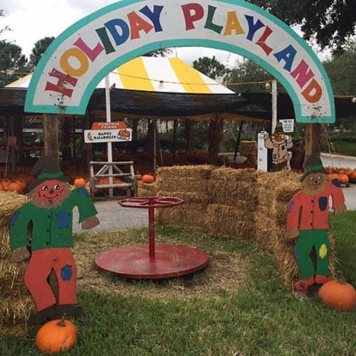 Colorful sign reading "Holiday Playland" above a festive area with hay bales, pumpkins, and playful scarecrow decorations.