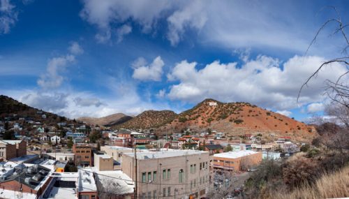 Panoramic view of a town nestled in hills, featuring colorful buildings and a blue sky with scattered clouds.