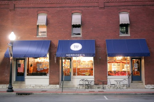 A brick storefront with blue awnings, featuring "Merridee's" signage and a display of items in the windows.