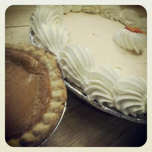 A slice of pumpkin pie next to a cream-topped dessert in a silver pie dish on a wooden surface.