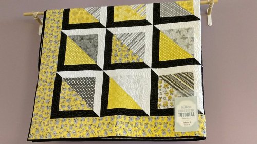 A colorful quilt featuring yellow, black, and gray patterns, displayed on a wall with a tutorial label attached.