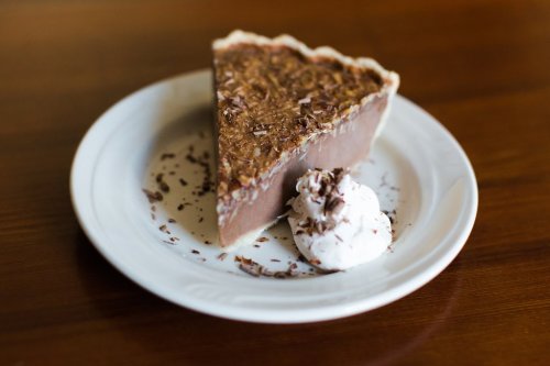 A slice of chocolate pie on a white plate, topped with whipped cream and chocolate shavings, on a wooden table.