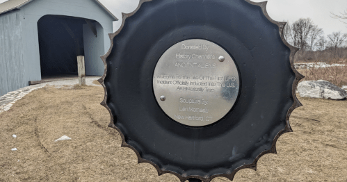 A metal plaque on a circular base, detailing a UFO incident, with a covered bridge in the background.