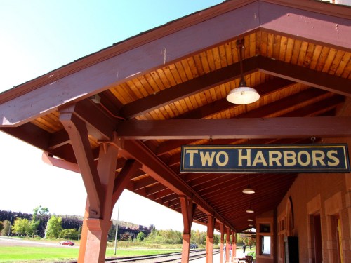 Sign reading "TWO HARBORS" on a train station platform with a wooden roof and clear blue sky in the background.