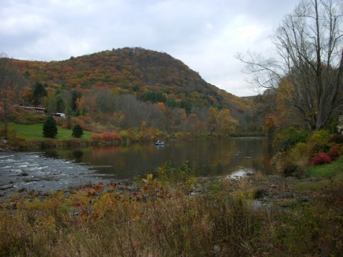 A serene river scene surrounded by autumn foliage and rolling hills under a cloudy sky. A small boat is on the water.