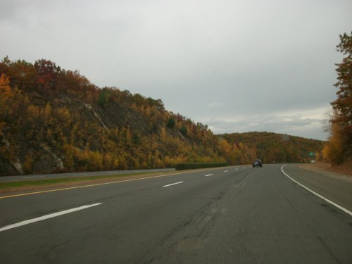 A scenic highway lined with autumn foliage on the hills, under a cloudy sky. A car drives along the road.