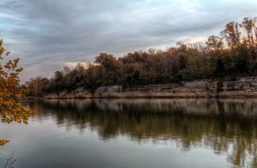 A calm river reflects a tree-lined shore under a cloudy sky, showcasing autumn colors on the trees.