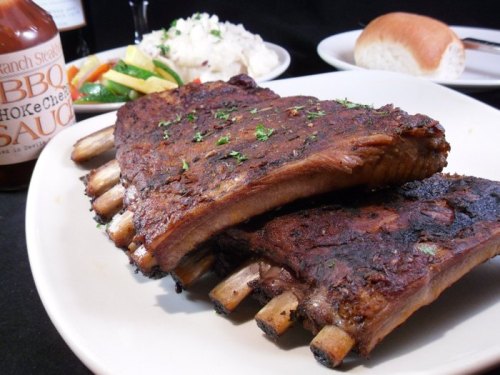 A plate of grilled ribs garnished with herbs, served with BBQ sauce, vegetables, mashed potatoes, and a bread roll.