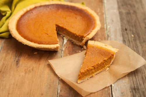 A pumpkin pie with a slice cut out, resting on a piece of parchment paper, on a rustic wooden table.
