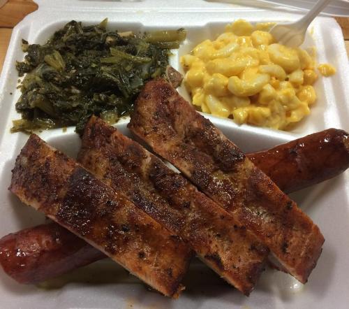 A takeout container with grilled sausage, smoked ribs, collard greens, and macaroni and cheese.