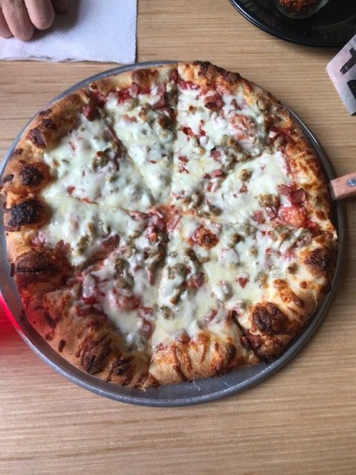 A round pizza with a golden crust, topped with melted cheese, chunks of meat, and green peppers, served on a metal tray.