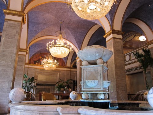 Elegant lobby featuring a marble fountain, chandeliers, and ornate arches with decorative murals.