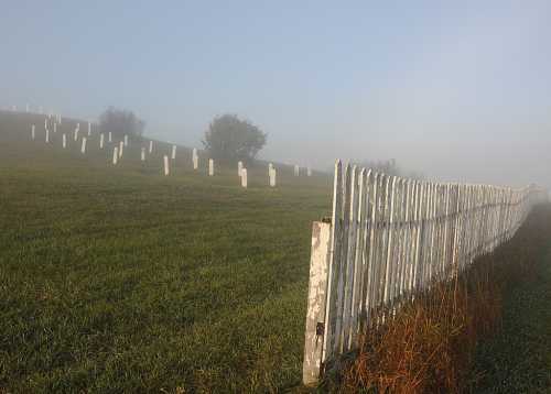 A foggy landscape featuring a white picket fence and gravestones on a grassy hill.