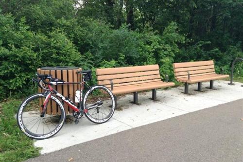 A red bicycle rests beside two wooden benches near a path, surrounded by lush greenery.