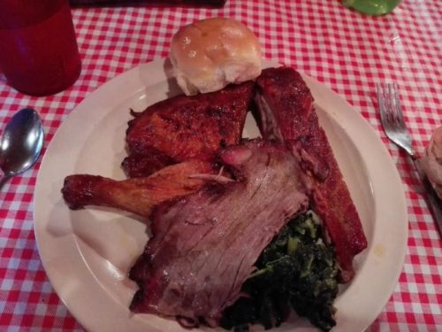 A plate of barbecue featuring ribs, chicken, brisket, collard greens, and a roll on a checkered tablecloth.