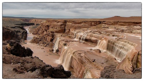 A scenic view of a canyon with cascading waterfalls and rocky cliffs under a cloudy sky.