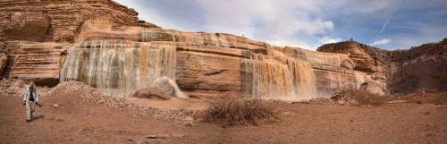 A panoramic view of a waterfall cascading over rocky cliffs in a desert landscape, with a person walking nearby.