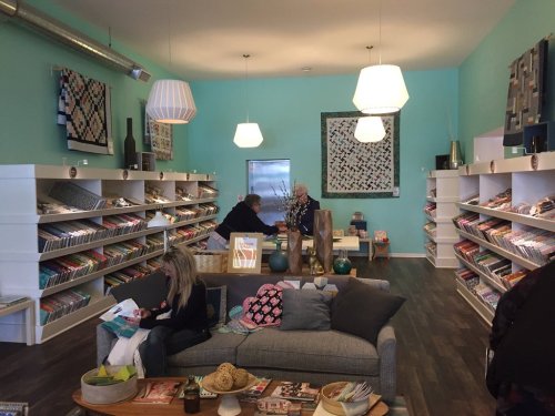 A cozy shop interior with teal walls, shelves of magazines, and a seating area with a sofa and decorative items.