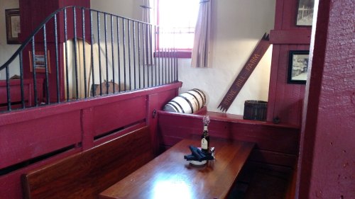 Cozy interior of a rustic room with a wooden table, a bottle, and barrels, featuring red walls and natural light.