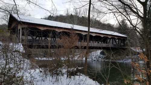 A wooden covered bridge spans a river, surrounded by trees and snow-covered ground in a winter landscape.