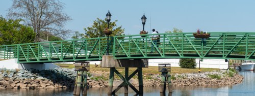 A green pedestrian bridge over a calm waterway, with two people fishing and decorative lamp posts along the path.