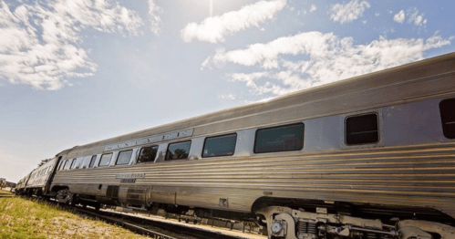 A silver train car on tracks under a blue sky with scattered clouds.
