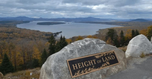 A scenic view from Height of Land, showcasing a lake surrounded by autumn foliage and distant mountains under a cloudy sky.