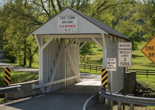 A covered bridge with a sign indicating weight limit and clearance, surrounded by greenery and a winding road.